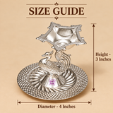 German Silver Peacock Pooja Diya size guide showing approximately 4 inch diameter and 3 inch height, with a raised decorative square diya bowl supported by an ornate peacock shaped centre and a circular embossed silver base, presented on a beige background with measurement arrows to help customers understand the compact dimensions for home mandir, daily pooja, Diwali lighting and gifting.