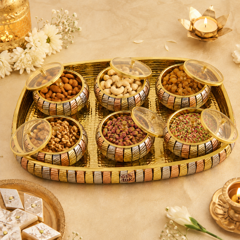 A decorative gold-toned serving tray holding six small patterned bowls filled with assorted nuts and dried fruits, arranged on a beige surface with flowers, candles, and a small plate of sweets around it.