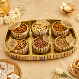 A decorative gold-toned serving tray holding six small patterned bowls filled with assorted nuts and dried fruits, arranged on a beige surface with flowers, candles, and a small plate of sweets around it.