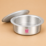 Aluminium Biryani Handi displayed with its matching circular lid resting at an angle behind the vessel, revealing the wide open cooking surface and deep round interior. The handi features a traditional hammered aluminium exterior with closely spaced circular impressions, a broad metallic rim, smooth inner surface and gently rounded bottom.