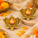 Two antique brass lotus diyas displayed in an elegant traditional puja setting on a warm beige marble surface, surrounded by yellow and orange marigold flowers, decorative brass accessories and festive fabric. Each diya features a golden lotus flower design, a central teardrop-shaped oil cavity and intricately textured petals, creating a traditional decorative arrangement for Diwali celebrations, daily worship and home mandir decoration.