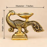 Size guide of a traditional antique brass peacock diya showing an overall horizontal measurement of 7 inches and a height of 4 inches. The decorative brass oil lamp features a sculpted peacock body with detailed feather engravings, curved neck, ornamental tail and a shallow oil-holding bowl positioned above its back. The structure rests on a rectangular pedestal with engraved decorative patterns. Measurement arrows highlight the compact dimensions of this traditional peacock deepam for home mandir worship, 