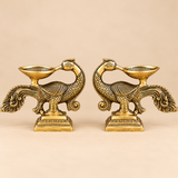 Pair of traditional antique brass peacock diyas displayed facing each other against a clean beige studio background. Each decorative brass oil lamp features a beautifully sculpted peacock with intricate feather engravings, curved neck, detailed wings, elaborate flowing tail and a shallow diya bowl mounted above the bird's body. The matching lamps rest on rectangular ornamental bases with detailed borders