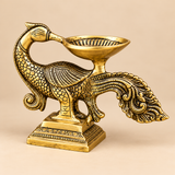 Angled view of an antique brass peacock diya showcasing its gracefully curved neck, finely engraved feather patterns, detailed wings and ornamental flowing tail. The traditional brass deepam features a shallow oil-holding bowl mounted above the peacock's body, supported by a decorative stem. The antique brass finish combines polished golden highlights with darker recessed detailing, enhancing the sculpted design. The lamp stands on a sturdy rectangular pedestal with engraved borders and is displayed against