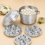 Ashtok aluminium idli steamer pot filled with freshly steamed idlis garnished with curry leaves, set alongside additional idli mold plates, a bowl of chutney, and a plate of idlis.