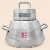 Front view of a sturdy silver aluminum tea brewing pot resting against a plain, light beige background. The pot features a tapered body, horizontal ridged decorative lines around its midsection, two thick side handles, a fitted cylindrical lid with a robust top handle, and a central magenta Ashtok logo watermark.