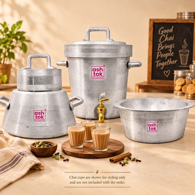 A styled product display featuring a set of three silver-colored aluminum vessels by Ashtok. On the left is a pot with side handles and a fitted lid; in the center stands a large tea container with side handles, a top lid, and a brass tap dispenser; on the right is a wide, open mixing bowl. In the foreground, three clear glasses filled with steaming hot chai sit on a round wooden tray alongside whole spices like cardamom pods, cloves, and cinnamon sticks. In the background, a small plant, a chalkboard sign 