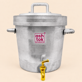 Frontal shot of a cylindrical silver aluminum tea dispenser vessel centered against a plain light beige backdrop. The vessel features a heavy lid with a top handle, two side handles, a polished brass spigot tap fixed at the bottom center, and the magenta Ashtok brand logo on the front.