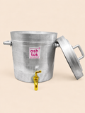 Side-angle view of a large, heavy-duty silver aluminum tea container set against a plain beige background. The container features side carrying handles, a golden brass tap dispenser near the bottom front, and a magenta Ashtok logo. The top lid with a top handle is shown unfastened and leaning against the right side of the vessel.
