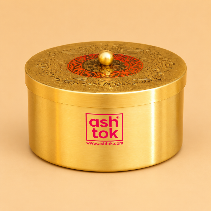 Product photo of a cylindrical brass decorative box by Ashtok set against a warm tan studio background. The container features a smooth, brushed gold finish on the body with the pink "ashtok" brand logo printed on the front. The top lid is intricately engraved with traditional patterns surrounding a central red circular ring with detailed accents and a small round gold knob handle in the middle.
