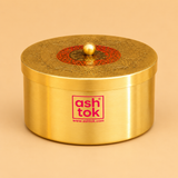 Product photo of a cylindrical brass decorative box by Ashtok set against a warm tan studio background. The container features a smooth, brushed gold finish on the body with the pink "ashtok" brand logo printed on the front. The top lid is intricately engraved with traditional patterns surrounding a central red circular ring with detailed accents and a small round gold knob handle in the middle.