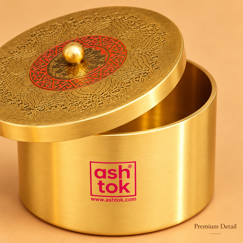 A close-up detail shot of the Ashtok brass container with its lid slightly off-center and tilted to reveal the interior rim. The image highlights the fine craftsmanship, showing the brushed metallic texture of the container side with the magenta logo, and the close details of the top lid’s intricate filigree engraving, red circular pattern, and central golden knob. "Premium Detail" text is subtly placed in the bottom right corner.