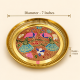 Product dimension diagram of the Ashtok round brass decorative tray against a warm studio background. Dimensional callout lines indicate a top outer diameter of 7 inches across the fluted rim and a side height of 1 inch. The center reveals intricate peacock Meenakari enamel artwork in pink, teal, blue, and red hues.