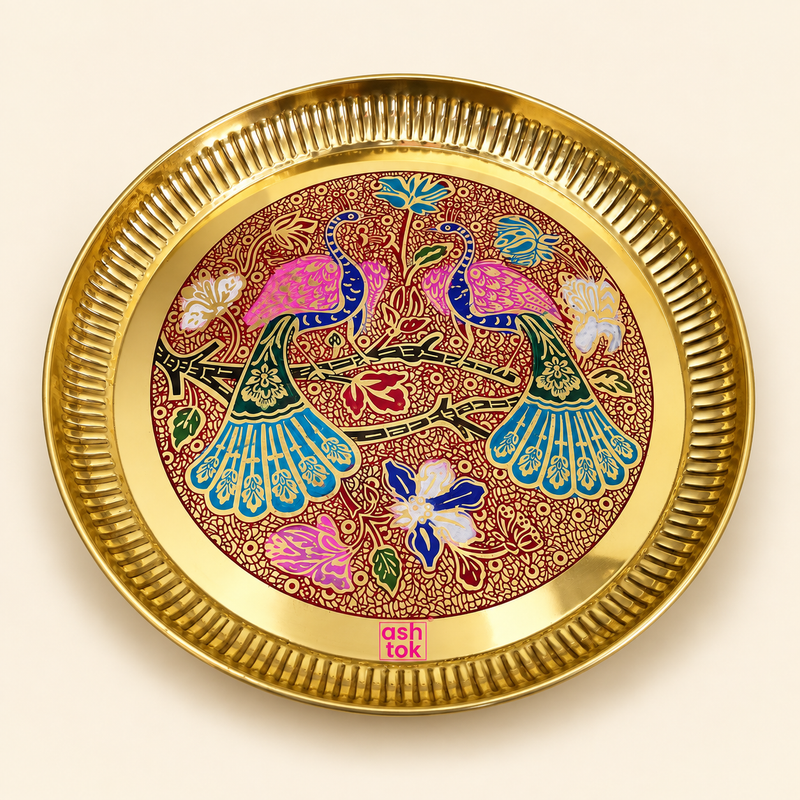 Top-down studio photograph of a round Ashtok brass decorative tray with a fluted rim. The interior of the tray features colorful Meenakari enamel artwork depicting two peacocks with pink, blue, and teal plumage perched amidst floral vines and blossoms against a textured red background, finished with a pink Ashtok logo at the bottom rim.