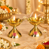 Festive lifestyle image of Ashtok brass diyas with lit flames placed in a decorative pooja setting with flowers and warm lighting on a beige background.