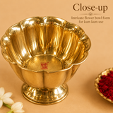 Close-up overhead shot of an empty Ashtok brand brass kumkum bowl designed in an intricate flower petal shape with a polished golden finish and a sturdy pedestal base, showcased next to a small dish of red kumkum powder and fresh white jasmine flowers.