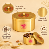 An infographic showcasing the features of the Ashtok brass decorative box. Callout circles highlight the "Decorative Engraved Lid" with its red circular accent, the "Brushed Metal Finish" of the body, "Ideal for Chocolates & Dry Fruits" with an illustration, and "Reusable Gift Box" represented by a recycling icon. In the foreground, an open matching container is filled with assorted chocolates, almonds, cashews, and pistachios.