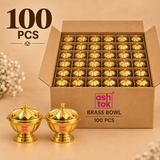 Product packaging photo of a 100-piece bulk set of Ashtok brass lidded kumkum bowls, displaying a large open cardboard box with grid dividers holding multiple golden containers, alongside two polished brass bowls placed in front.
