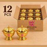 Product packaging showcase for a 12-piece set of Ashtok lidded brass kumkum bowls, showing an open cardboard storage box with partitioned slots holding brass containers, with two golden lidded bowls positioned in the foreground.