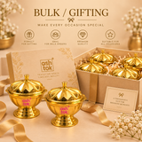 Bulk gifting display for Ashtok brass lidded kumkum boxes, showing two polished brass containers in the foreground alongside an open cardboard gift box filled with paper shredding and four lidded brass containers. The scene includes golden satin ribbons, white baby's breath flowers, and a gift card reading "A thoughtful gift for brighter moments."