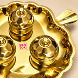 Close-up detail of the ASHTOK brass leaf shaped kumkum box set showing the highly polished golden brass surface, scalloped leaf-shaped tray rim, decorative leaf handle and three matching round containers with faceted dome-style lids and small knobs. The close view highlights the smooth reflective finish, lid detailing, metal craftsmanship and pink ASHTOK logo, presented against a clean warm beige background.
