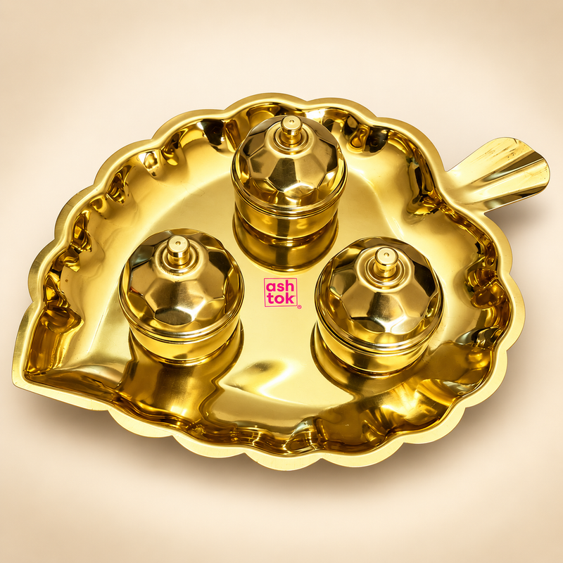 ASHTOK brass leaf shaped kumkum box gift set featuring a polished golden brass leaf tray with decorative scalloped edges, a short leaf-style handle and three matching round kumkum containers with fitted faceted brass lids and small top knobs. The complete set is displayed on a clean warm beige studio background, clearly showing the reflective brass finish, traditional design and pink ASHTOK branding, suitable for pooja use, festive gifting, housewarming gifts, wedding gifts and return gifts.