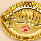 Detailed close-up image of the Ashtok brass plate shankh shape showing the fine craftsmanship and premium finish of the product. The close-up highlights the shiny golden brass surface, raised embossed lines inside the plate, scalloped edge detailing and the graceful shankh-inspired contour of the plate. The original pink Ashtok logo is clearly visible in the inner base area, and the polished reflective finish gives the product a rich and luxurious appearance. This close-up is useful for customers who want t