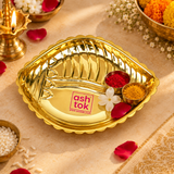 Lifestyle image of the Ashtok brass plate shankh shape styled in a festive and traditional Indian pooja setting. The polished golden brass plate is placed on a warm beige surface and is accompanied by small bowls of haldi and kumkum, flower petals, white flowers, a decorative brass diya and festive floral accents in the background. The product retains its elegant shankh-inspired form, scalloped rim, embossed pattern details and reflective brass shine, while the original pink Ashtok logo remains visible. Thi