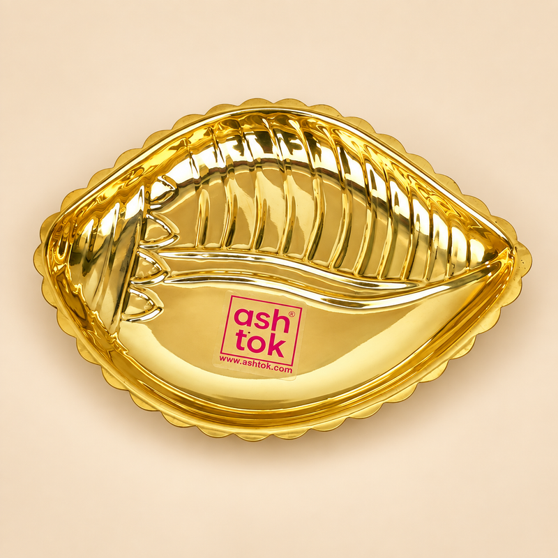Ashtok brass plate shankh shape displayed as a single premium decorative brass plate on a clean warm beige studio background. The product features a polished golden brass finish, a distinctive shankh-inspired shape, a scalloped outer edge and raised embossed detailing inside the plate that gives it an elegant traditional appearance. The plate has a reflective glossy surface and clearly shows the original pink Ashtok branding placed at the center area. This brass shankh shape plate measures approximately 5.5