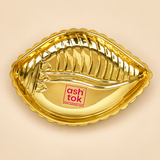 Ashtok brass plate shankh shape displayed as a single premium decorative brass plate on a clean warm beige studio background. The product features a polished golden brass finish, a distinctive shankh-inspired shape, a scalloped outer edge and raised embossed detailing inside the plate that gives it an elegant traditional appearance. The plate has a reflective glossy surface and clearly shows the original pink Ashtok branding placed at the center area. This brass shankh shape plate measures approximately 5.5