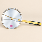 Top-down view of an inner-lid pressure cooker cover from the brand Ashtok, isolated against a solid beige background. The lid features a round, shiny metallic inner plate with a soft brushed finish, stamped with a red "ashtok" logo near the bottom left edge. At the center sits a raised gold-colored brass vent pipe surrounded by two slight structural ridges extending outward across the lid. To the right of the center vent pipe, a round brass safety valve with a black seal is embedded in the surface. Extendin