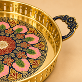 Close-up detail of ASHTOK brass round serving tray highlighting the polished golden brass finish, intricate perforated side rim, decorative blue and gold handle craftsmanship and vibrant multicolor floral center artwork for premium serving and display.