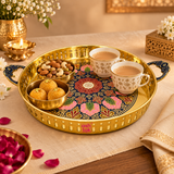 ASHTOK brass round serving tray displayed in a premium festive setting with tea cups, dry fruits and sweets, featuring a colorful floral center pattern, polished golden brass body, perforated rim and decorative handles for elegant home serving.