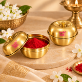 Ashtok Brass Sindoor Box shown in an elegant traditional lifestyle setting with one glossy golden brass container open and filled with bright red sindoor or kumkum and another matching brass box displayed closed with its fitted lid. The polished brass containers feature the original pink Ashtok logo and are styled on a warm beige surface with white flowers, green leaves, festive fabric and a traditional brass lamp in the softly blurred background. This lifestyle image demonstrates how the compact brass sind