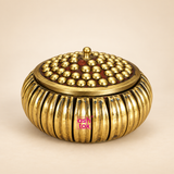 Close-up view of ASHTOK brass sindoor kum kum box with lid open, showing the polished brass interior, decorative beaded lid and traditional ribbed outer body on a beige background.