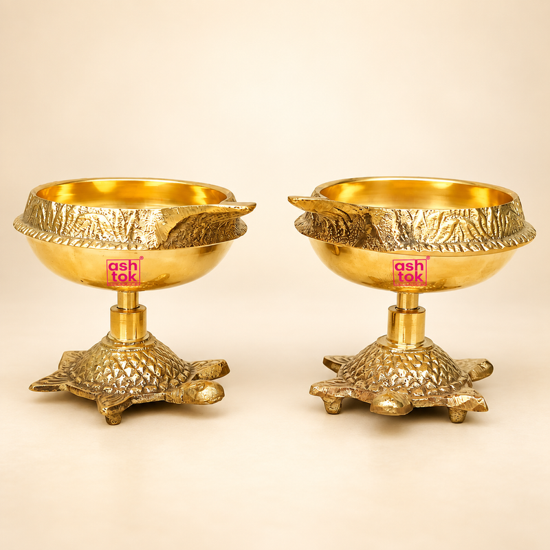 Set of two polished brass diyas with decorative turtle-shaped bases, displayed on a warm beige background, ideal for pooja decor, festive use, and traditional home lighting.