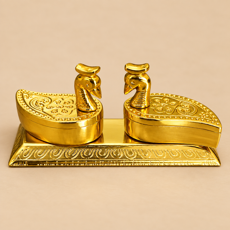 Decorative brass twin peacock haldi kumkum box displayed as a single premium traditional storage piece on a clean warm beige studio background. The product features two beautifully shaped compartments with curved peacock-inspired lids, detailed embossed floral and scrollwork patterns on the top, smooth rounded sides, polished golden brass finish and a matching rectangular ornamental base. The peacock head knobs create a graceful symmetrical look, making the piece suitable for storing haldi and kumkum separa