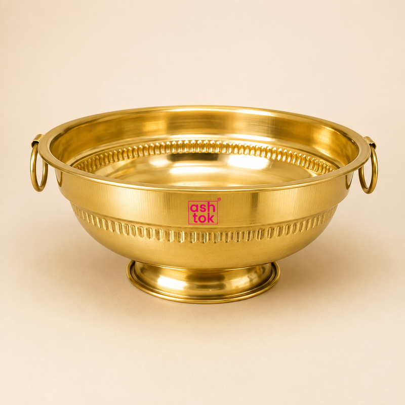 Ashtok traditional brass urli bowl featuring a raised pedestal base, side ring handles, and decorative ribbed detailing.