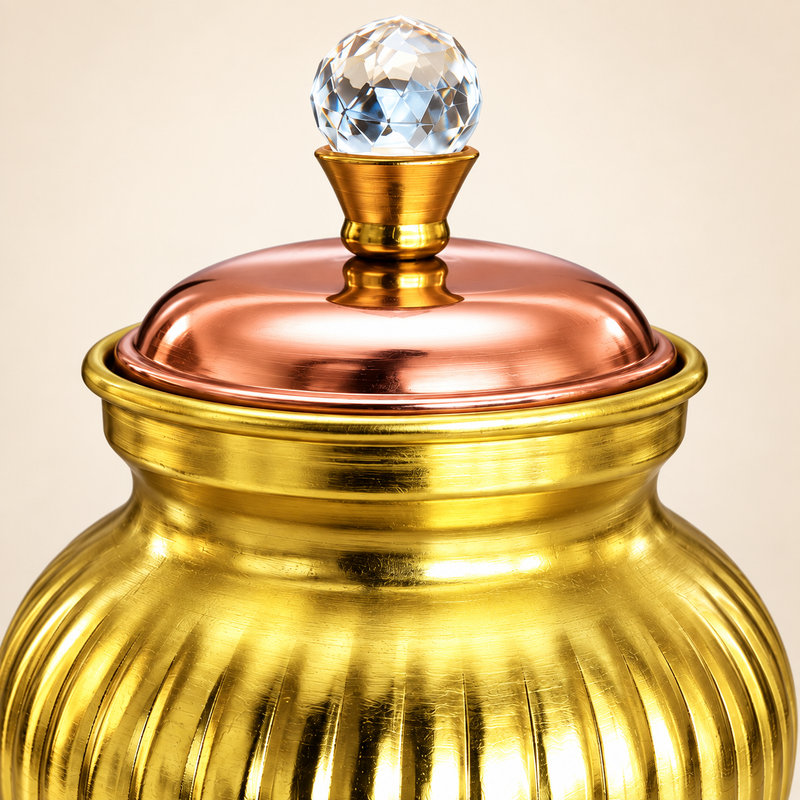 Ashtok chocolate box surai gift item in a clean beige studio setting, featuring a decorative round brass-finish storage jar with vertical ribbed detailing, a warm copper-toned lid, and a faceted crystal-style knob on top. The product is shown as a standalone hero image with soft lighting and a neat shadow, clearly highlighting the elegant shape, polished metallic finish, rounded body, and premium handcrafted appearance. This decorative gift box style surai is ideal for festive gifting, storing chocolates or