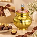 Detailed product showcase of the Ashtok chocolate box surai highlighting the elegant craftsmanship, decorative shape, and refined metallic finish. The image emphasizes the rounded ribbed body in a warm brass-gold tone, the smooth copper-toned lid, and the crystal-style top knob that gives the gift item a premium and ornamental appearance. Set against a beige background, the composition focuses on product finish, symmetry, design detailing, and overall gifting appeal. This image is useful for customers who w