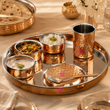 Ashtok hammered copper dinner set arranged with cooked Indian food, rice, curries, yogurt, and a copper spoon on an elegant dining setup.