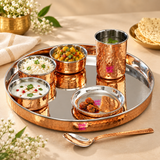 High-angle lifestyle product shot of a hammered copper dinner thali set by Ashtok placed on a light beige surface. The set features a large round copper tray with a stainless steel interior lining. Arranged on the tray are three small copper bowls containing cooked white rice garnished with herbs, a yellow potato and pea curry, and white curd with garnishes. A small copper dish holds pickle pieces, and a hammered copper glass filled with water sits in the upper right corner of the plate. A matching copper s
