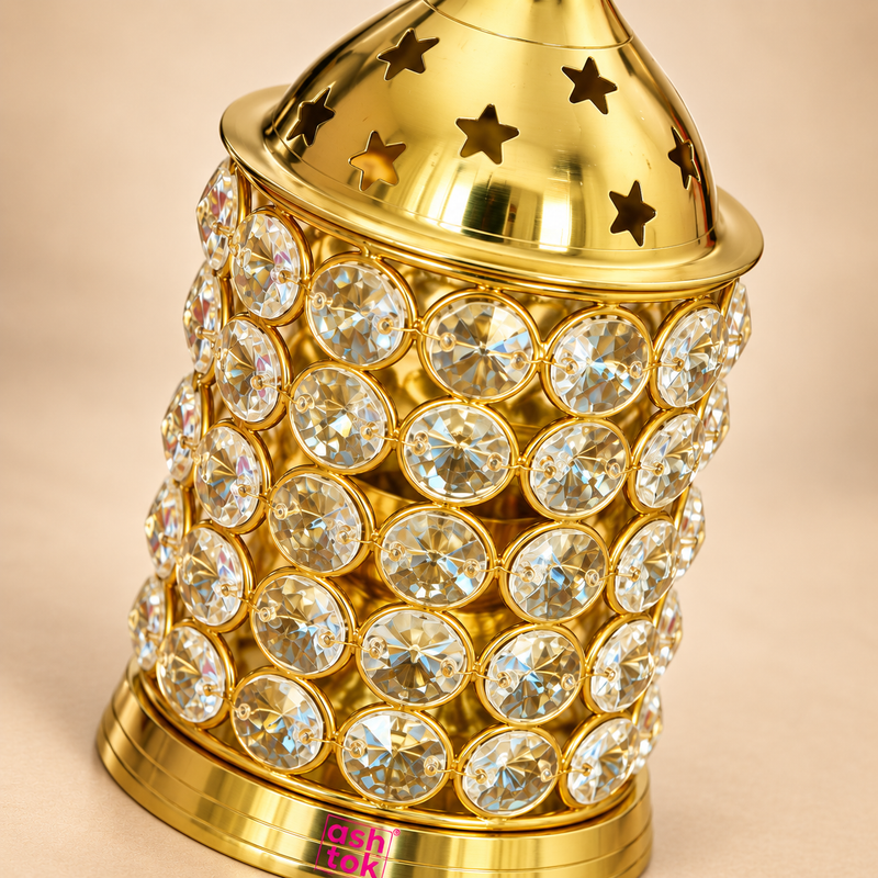 Close-up detail image of the ASHTOK Crystal Diya Brass focusing on the upper and middle sections of the product. The photograph clearly shows the polished brass dome with star-shaped cutouts, the fine reflective metal finish, and the crystal-studded cylindrical body made of round clear stones set in a metal frame. This detailed close-up highlights the craftsmanship, premium materials and decorative appeal of the diya, making the sparkle, texture and finishing clearly visible.
