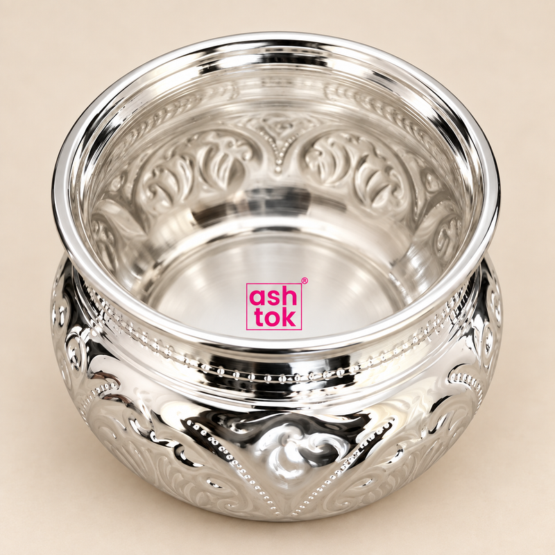 Detailed close-up view of the Ashtok German Silver Chopala Haldi Kumkum Box shown from a slightly elevated top angle on a clean beige studio background. The close-up highlights the smooth polished interior, wide flared silver rim, reflective German silver finish, intricate embossed floral and scroll patterns around the inner and outer body, fine beaded decorative detailing and the compact rounded form of the bowl. The original pink Ashtok logo is clearly visible, while the close framing helps customers insp