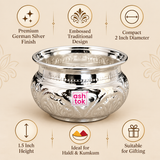Ashtok German Silver Chopala features image highlighting the premium German silver finish, embossed traditional design, compact 2 inch diameter, 1.5 inch height, suitability for haldi and kumkum storage and use as an elegant gifting item. The polished silver-finish bowl is displayed prominently with its wide flared rim, rounded pot-style body, decorative embossed floral detailing and original pink Ashtok logo. Feature icons surrounding the product explain the compact size, traditional craftsmanship and gift