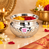 Ashtok German Silver Chopala Haldi Kumkum Box shown in an elegant traditional festive lifestyle setting with bright yellow haldi and deep red kumkum placed inside the polished decorative bowl. The German silver chopala features a wide shiny rim, rounded embossed body, detailed floral ornamentation and clearly visible pink Ashtok logo, styled on a warm beige surface with flowers, traditional brass décor, soft festive lighting and decorative fabric in the surroundings. This image demonstrates how the chopala 