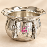 Close-up detail image of the ASHTOK German Silver handi and sweet bowl showing the reflective hammered silver body, wide scalloped rim, twisted rope-style upper band, detailed hanging tassel ornaments and bright pink ASHTOK logo. The close view emphasizes the polished finish, decorative craftsmanship and elegant contours of the bowl against a clean warm beige background.