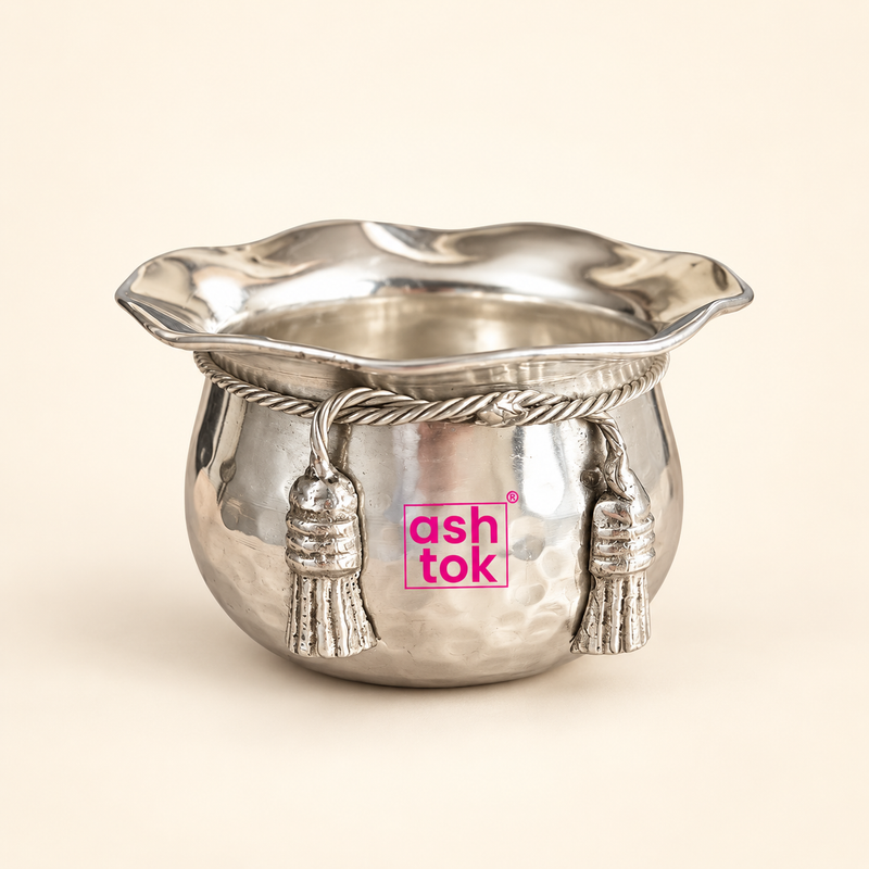 ASHTOK German Silver handi and sweet bowl displayed as a single premium serving bowl on a clean warm beige background, featuring a rounded hammered silver-finish body, wide flared scalloped rim, twisted rope-style decorative band and two hanging tassel ornaments on the front. The polished reflective finish and pink ASHTOK branding are clearly visible, making the bowl suitable for serving sweets, dry fruits, festive treats, gifting, return gifts and decorative table presentation.