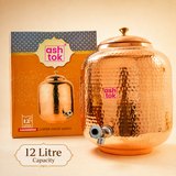 Ashtok 12-litre hammered copper water matka dispenser featuring a chrome tap and lid, next to its official product box.