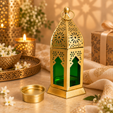 Lifestyle image of the Ashtok Moroccan Lantern Candle Holder displayed in an elegant warm-toned décor setting with decorative flowers, candlelight, festive accents and premium fabric styling. The gold Moroccan lantern with green glass panels is placed prominently on a tabletop with its removable candle cup beside it, showing how the product can be used beautifully in home décor settings, festive table decoration, ambient corner styling, living room decoration, Ramadan or celebration décor, gifting displays 